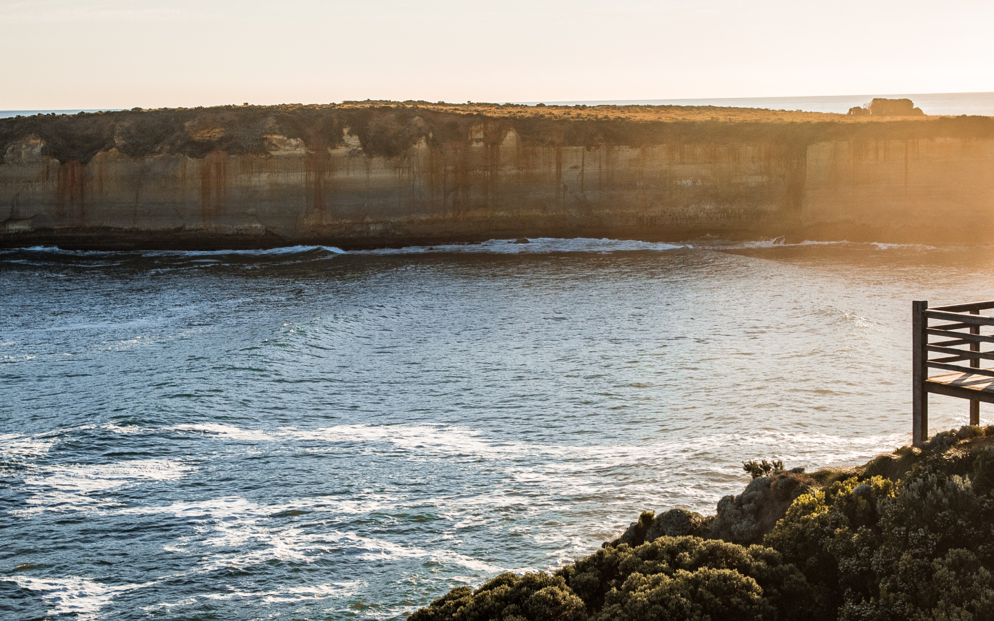 A large cliff overlooks a rough body of water with ocean in the distance and foliage and a wooden deck in the foreground.