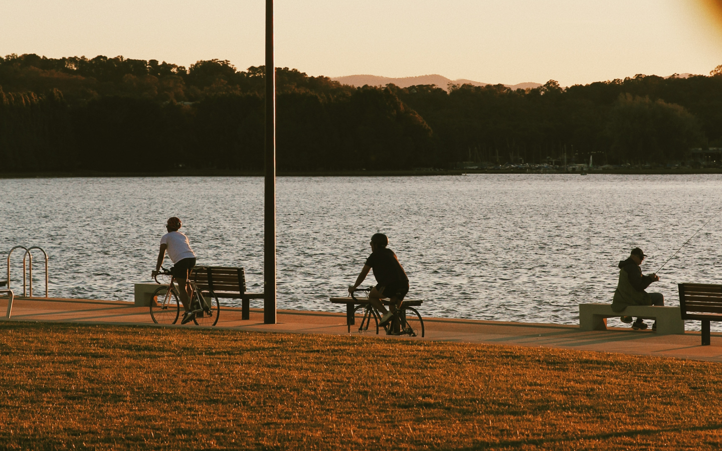 Two people on bicycles ride on a path next to a man fishing in a body of water.