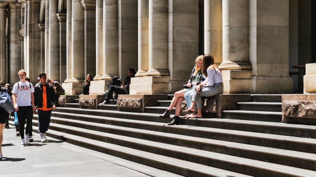 Two conversing women seated on steps in a city environment with passers by.