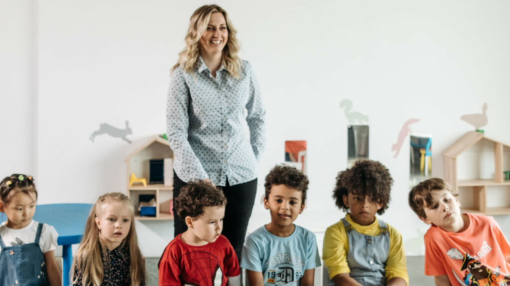 Female teacher looking happy, standing in classroom with group of six children in foreground.