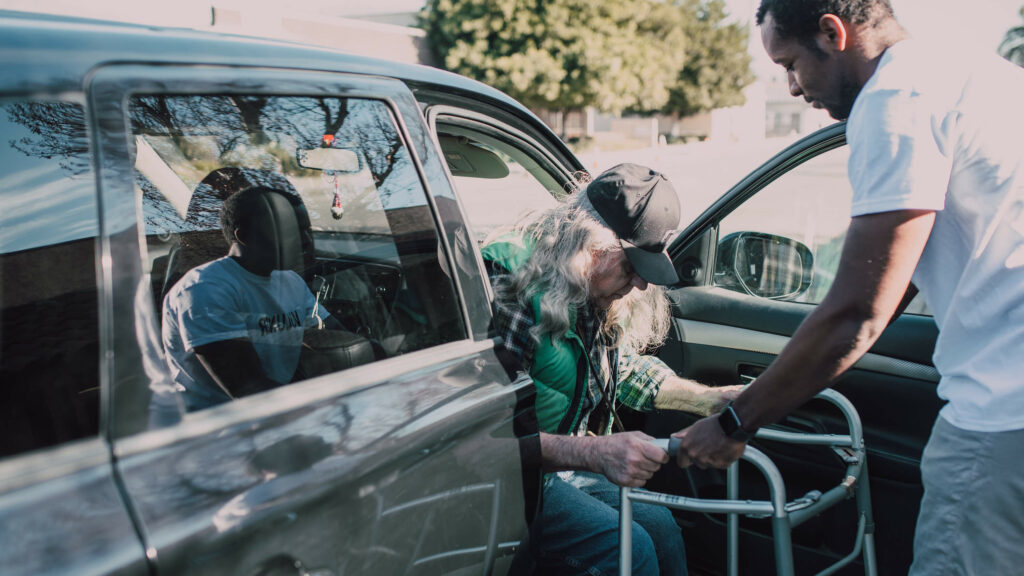 Photo of man helping an elderly person on walking frame out of a car.