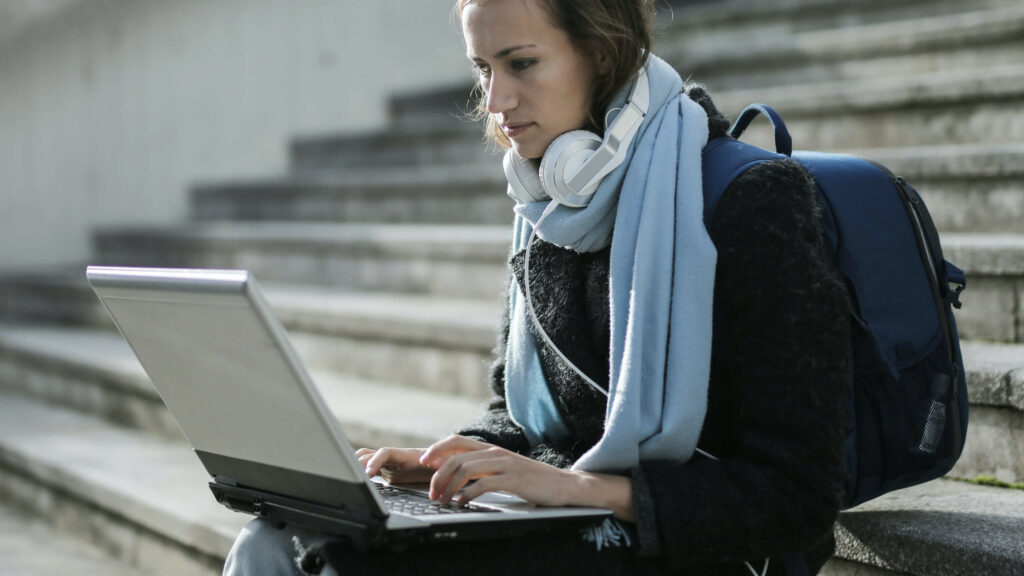 A woman with scarf and headphones is typing on a laptop computer while sitting on steps.