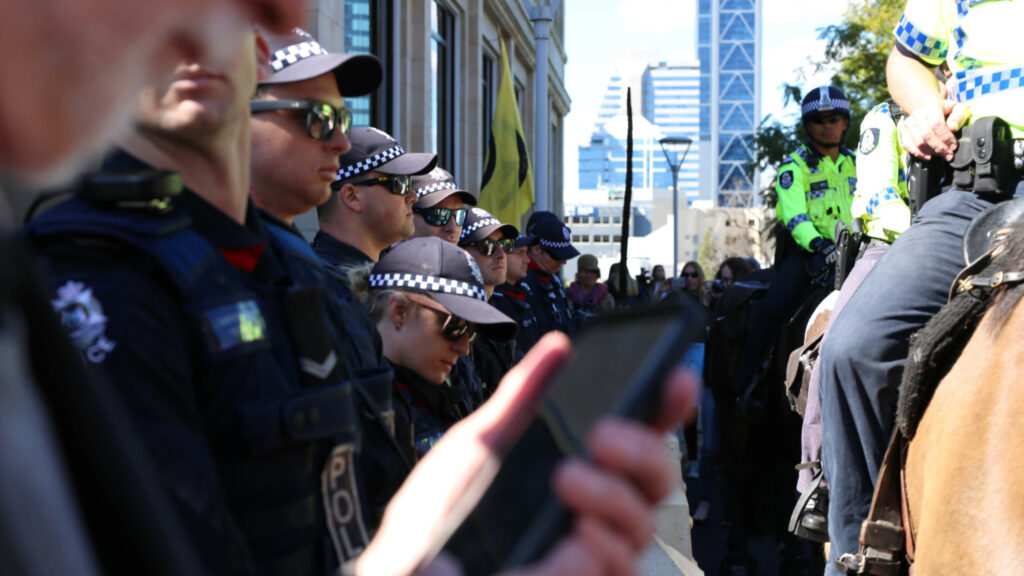 Photo of NSW police officers in uniform standing in a row with two police officers in uniform riding horses in large crowd.