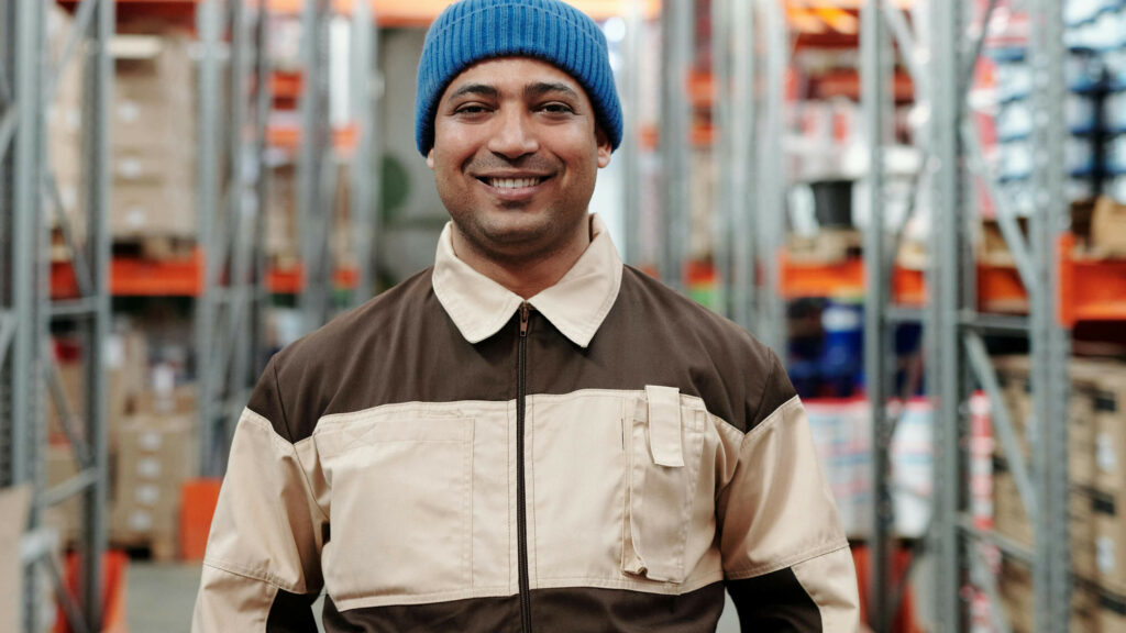 Photo of man looking to camera in factory shelving with blue beanie and brown jumper.
