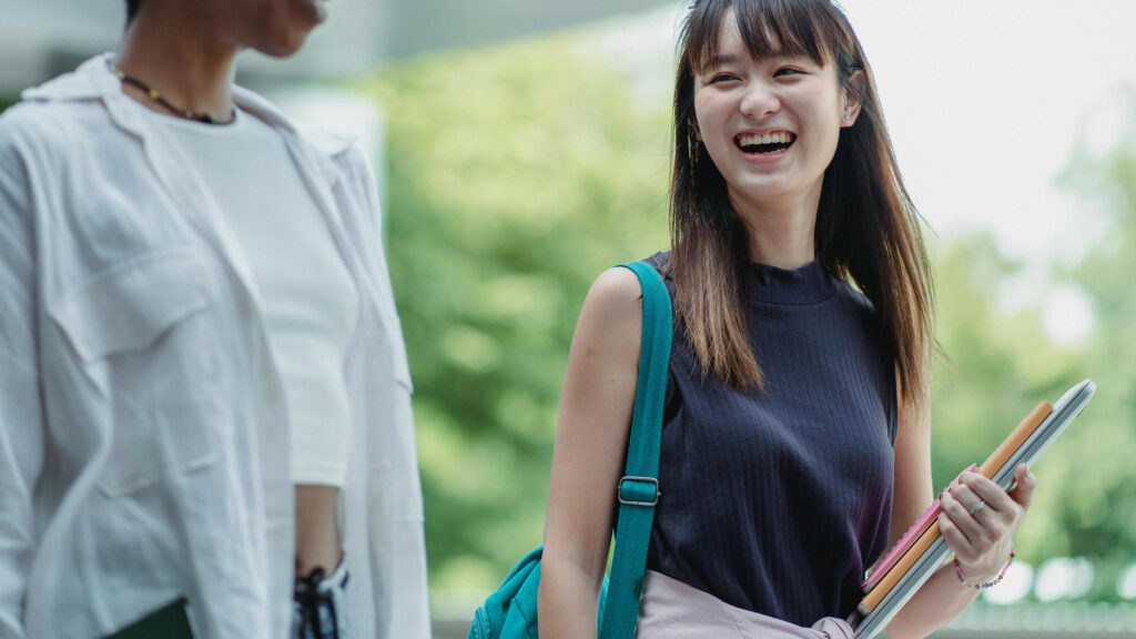 Two women outdoors, one carrying a shoulder bag, books and a laptop.