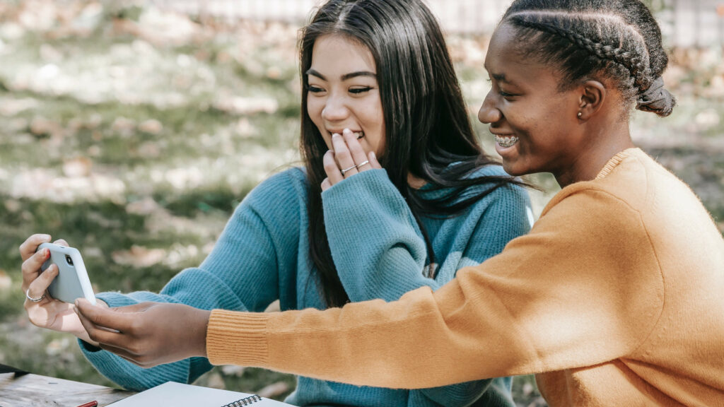 Two seated women are laughing to a smart phone.