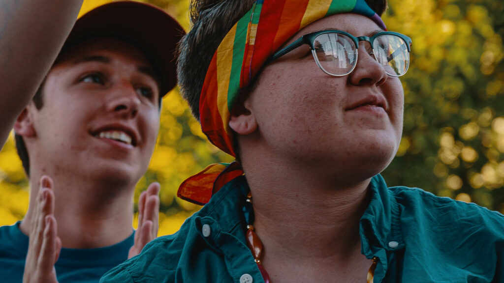 Photograph of two people looking happy, one is clapping and the other wears glasses and a rainbow bandanna.