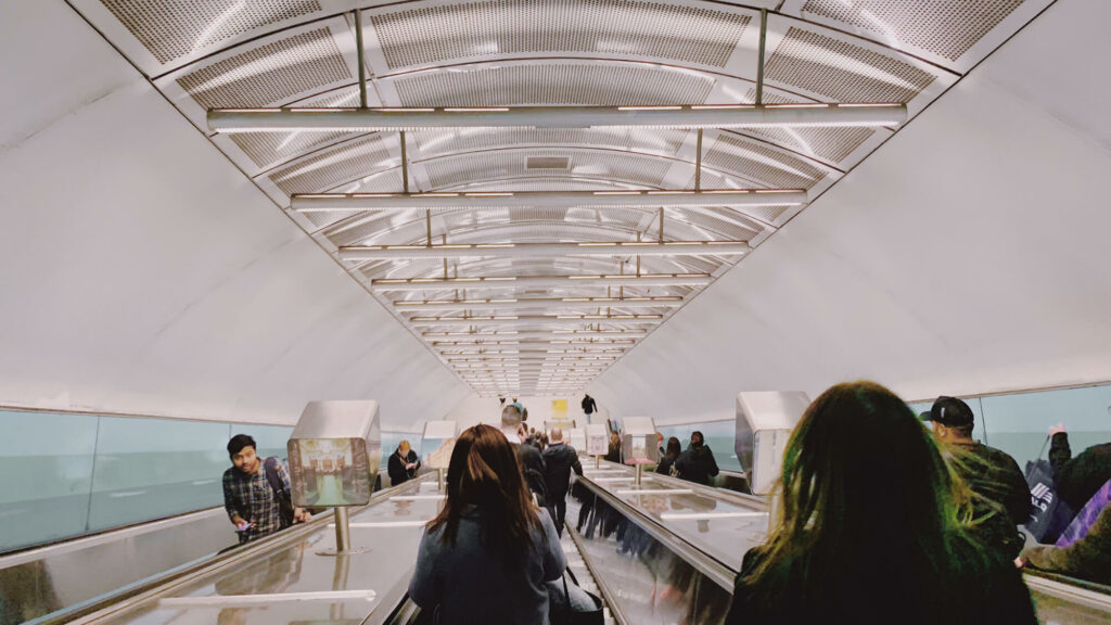 Wide photograph of people going up and down silver escalators with a white roof.