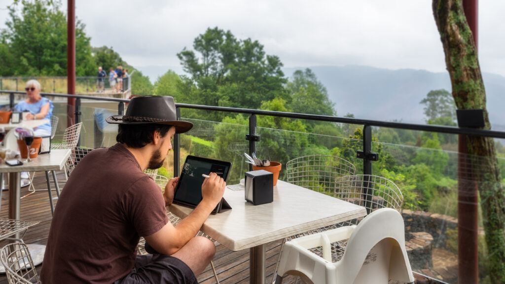 Young man drawing using his laptop, while relaxing at a cafe terrace after rain at Mount Tomah Botanic Garden in Blue Mountains National Park, NSW, Australia.
